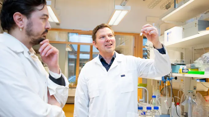 Two researchers in lab coats discuss while one holds up a test tube.