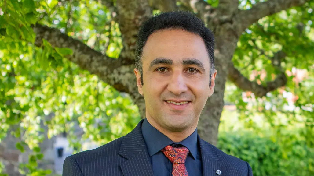 A person with dark hair, wearing a dark blue suit, looks into the camera.