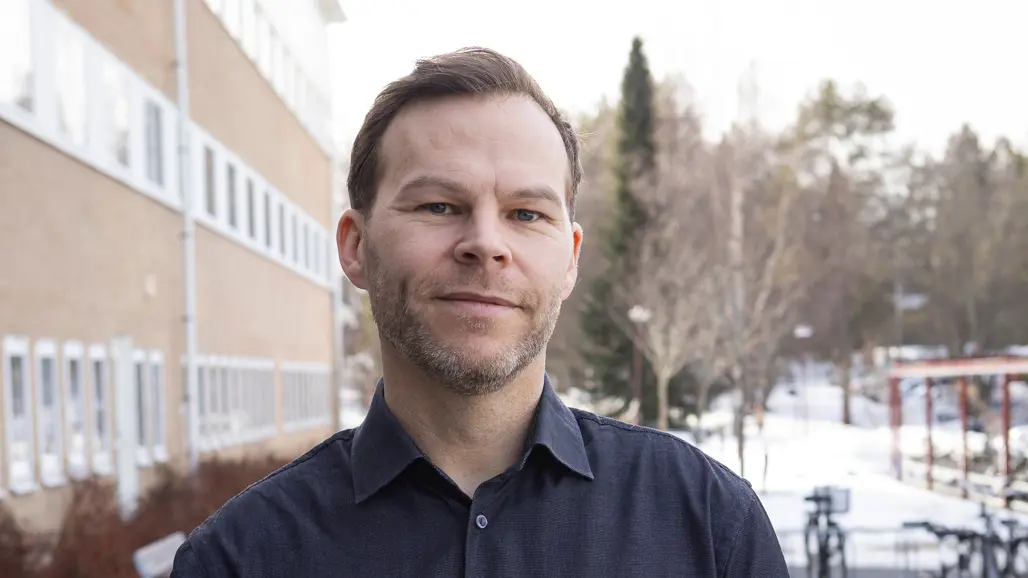 En person i mörk skjorta står utanför ett hus på vintern och tittar i kameran.