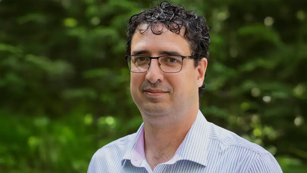 A man with glasses and dark short curly hair looks into the camera. Green lush background.