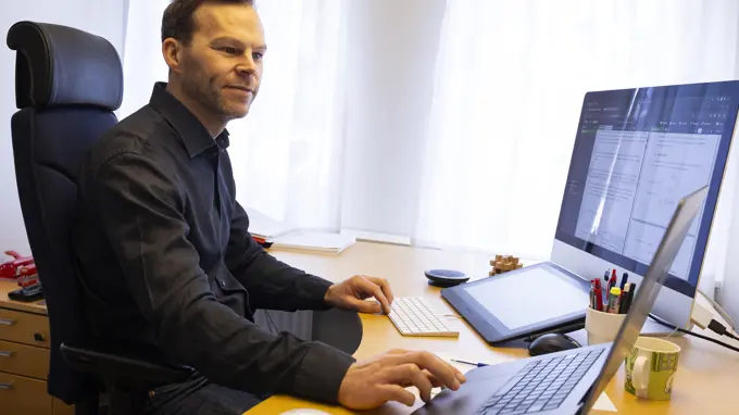 En man i svart skjorta sitter framför ett skrivbord med en dator och en pekplatta.