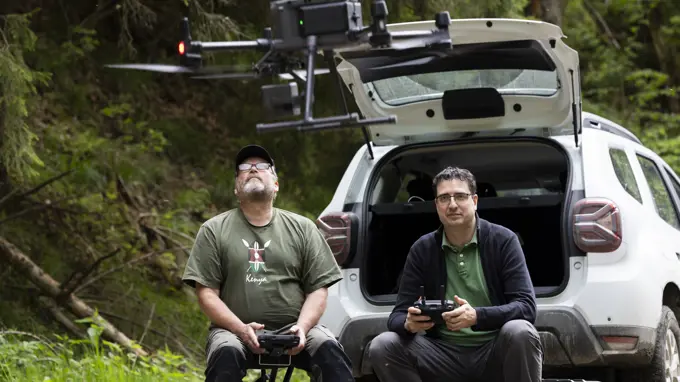 Two persons sitting in open trunk of a car, holding remote controls, one of the persons are looking at a drone hovering in the air.