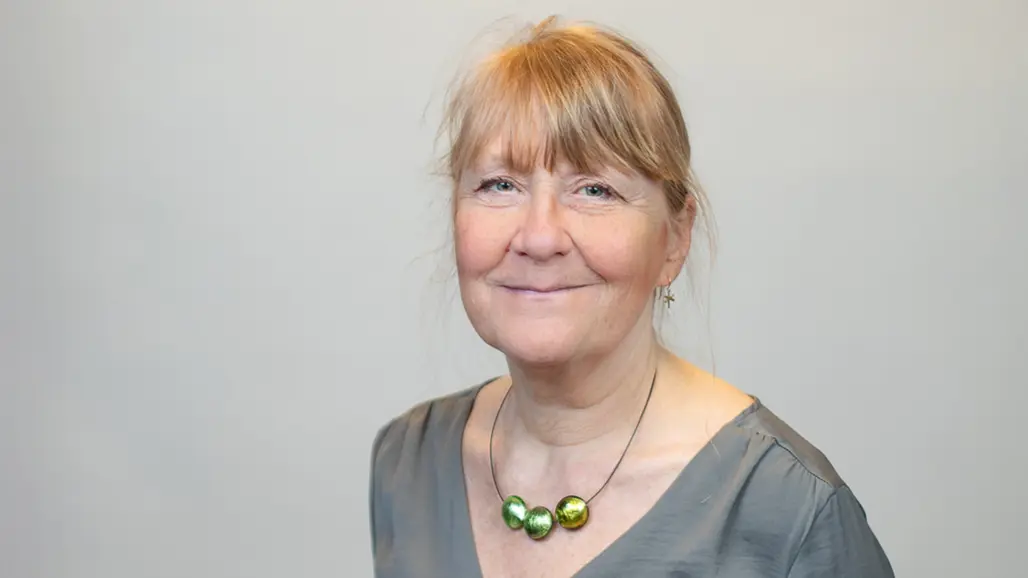 A woman with reddish blonde hair and a necklace with three big green pearls smiles into the camera.