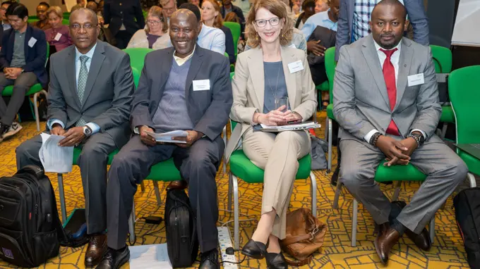 A woman and three men, all of them wearing suits and nametags, sit on green chairs in a row, smiling into the camera.