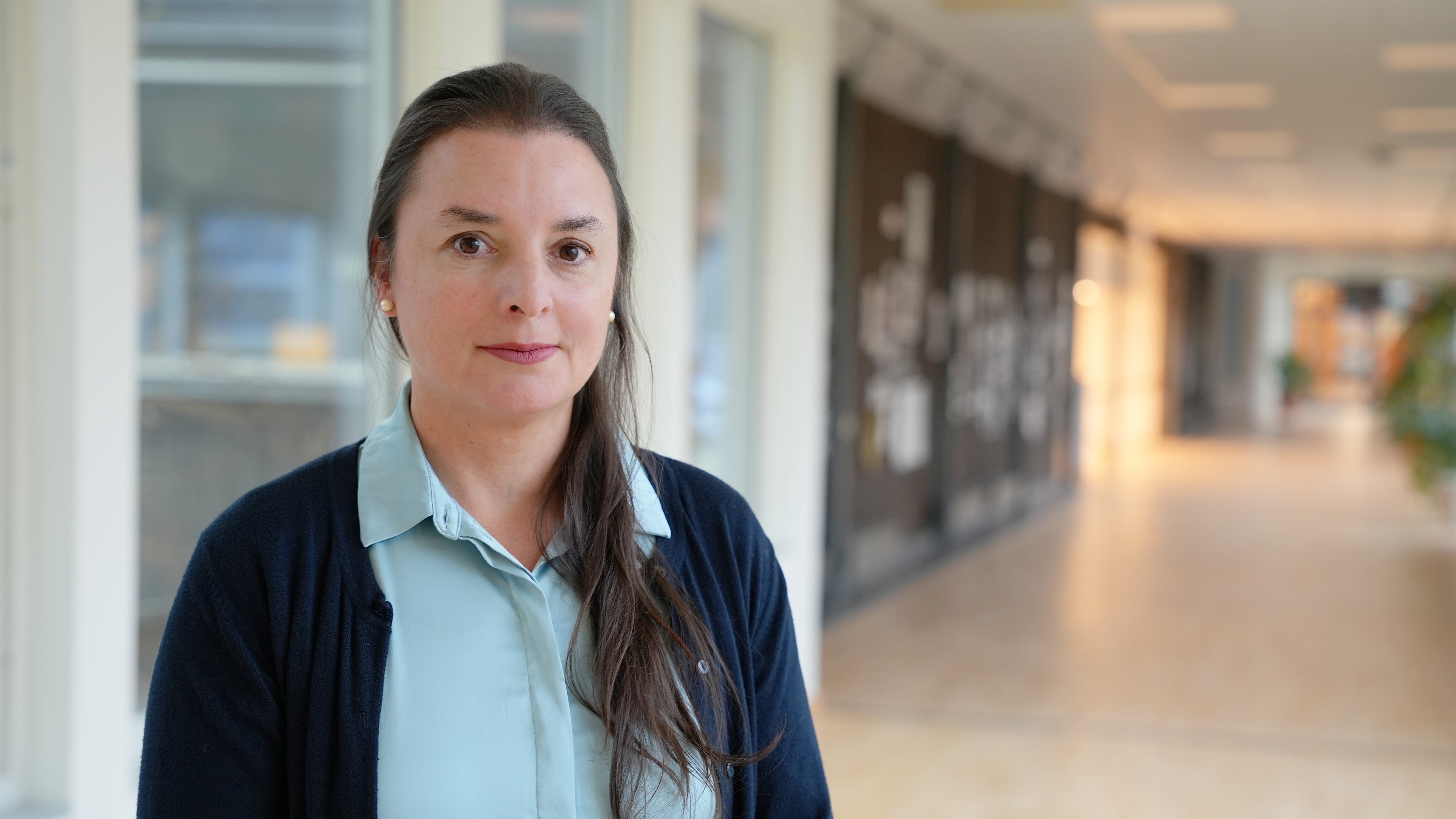 A woman with dark brown hair, wearing a blue shirt looks into the camera. 