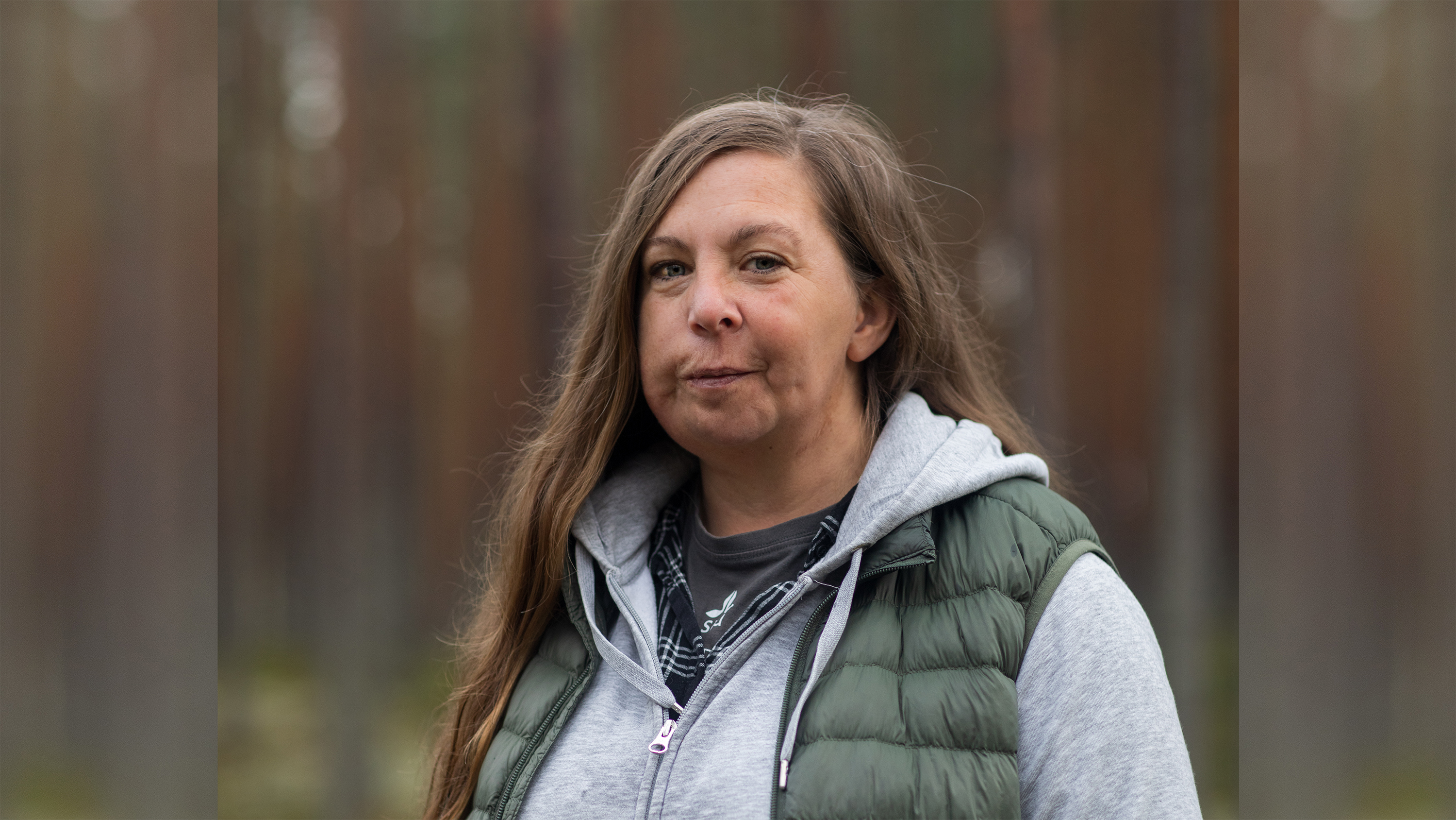 En kvinna med långt brunt hår och grön täckväst står i skogen.