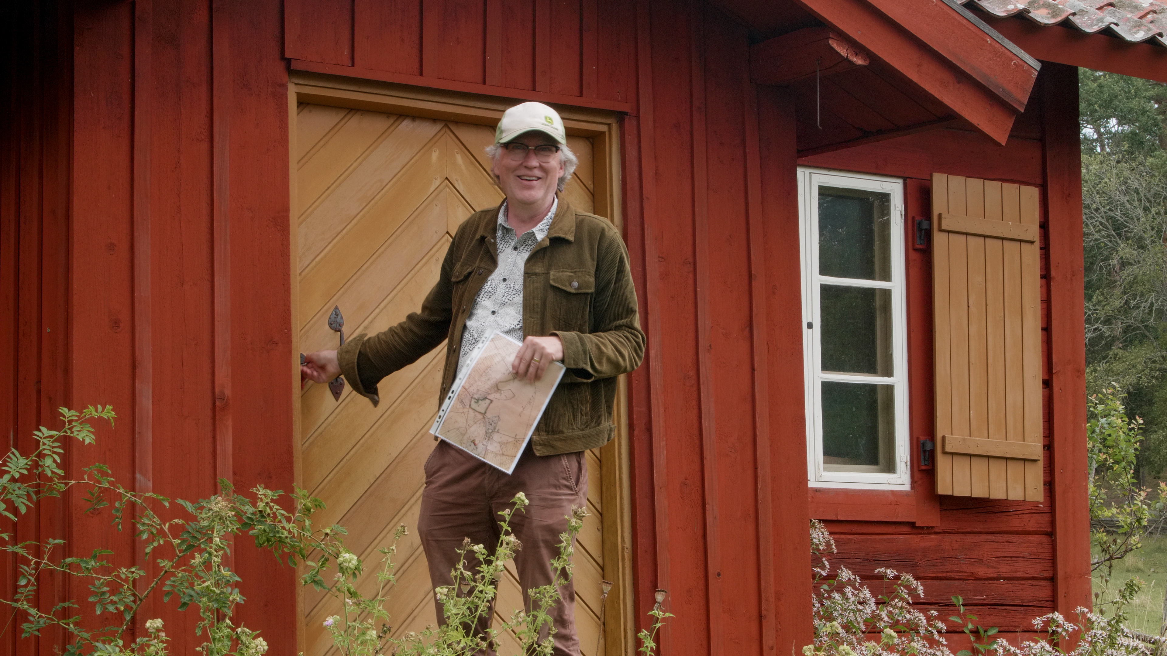 En man i keps står på trappen till ett gammalt hus, vänd mot kameran. 