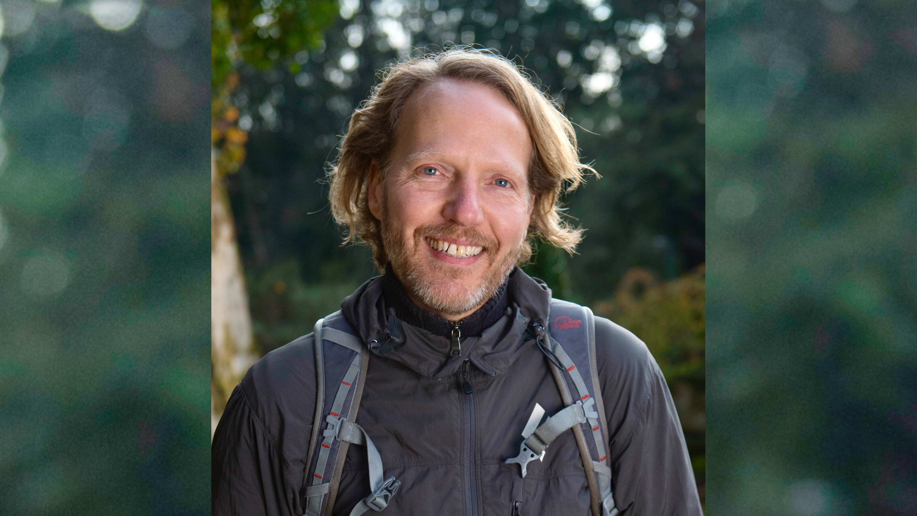 En man med blont långt hår står i friluftskläder och ryggsäck i skogen. 