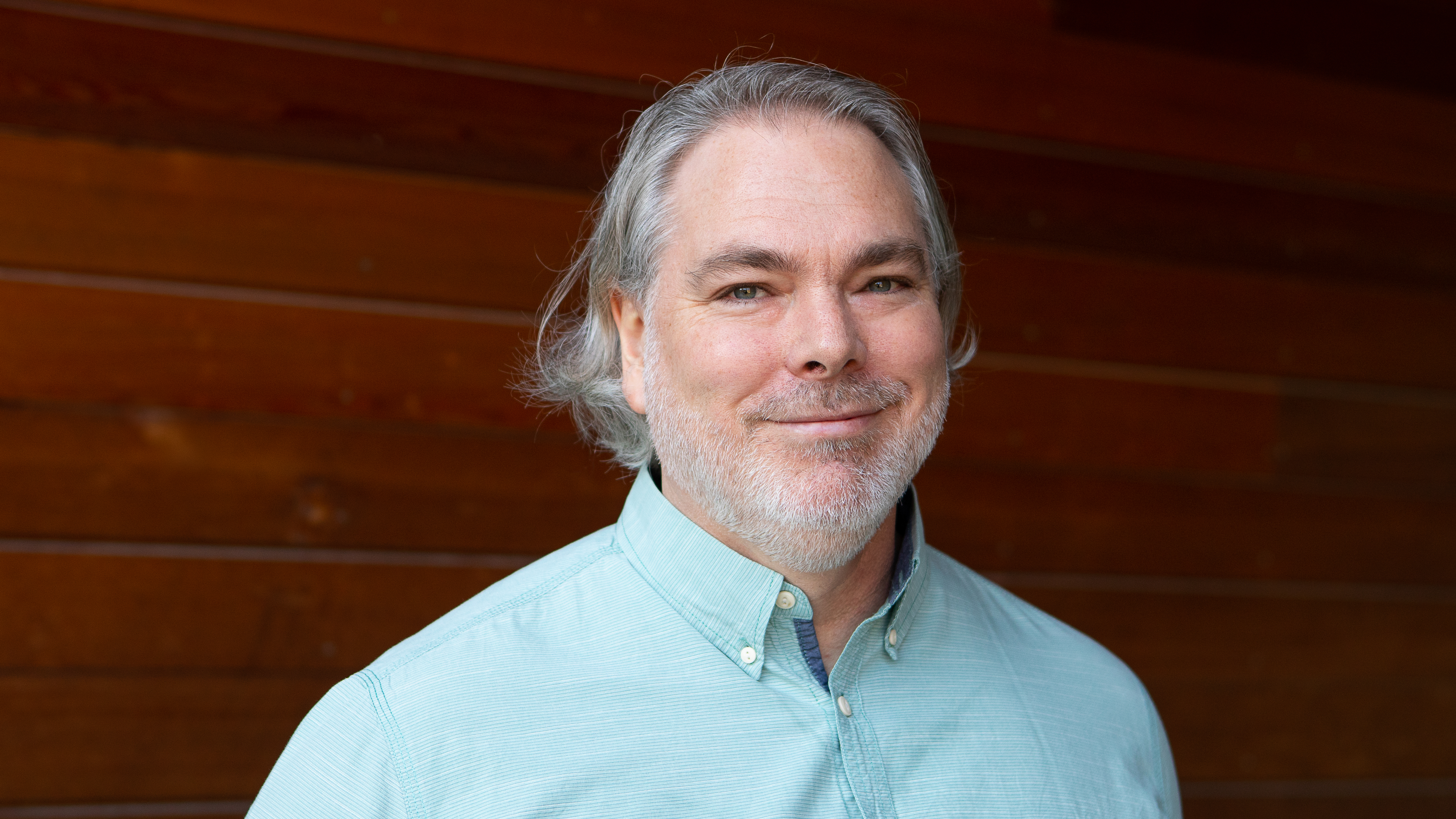 En leende man i ljus skjorta och långt hår står framför en trävägg. 