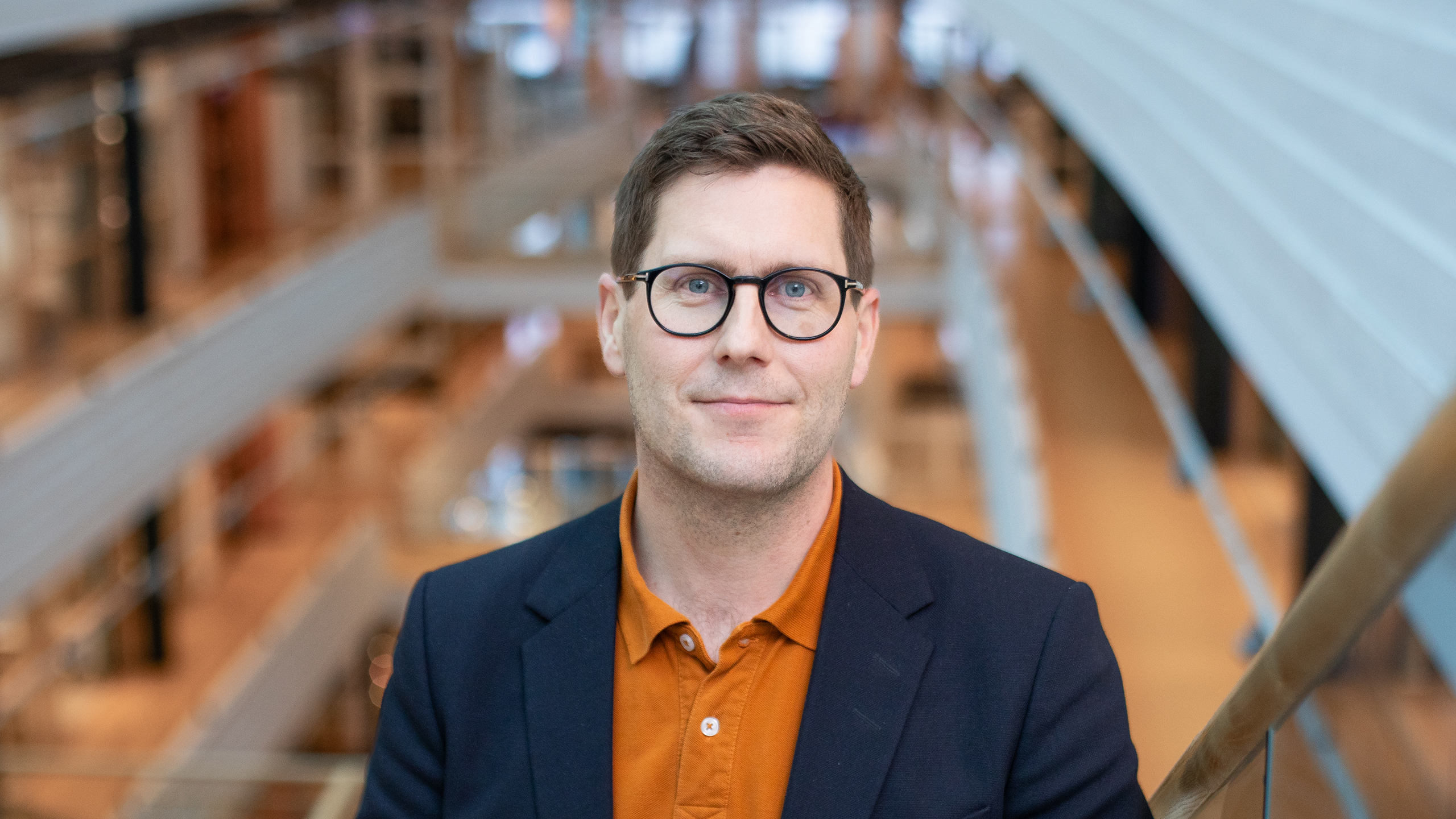 En man i glasögon, kavaj och orange pikétröja tittar leende in i kameran.