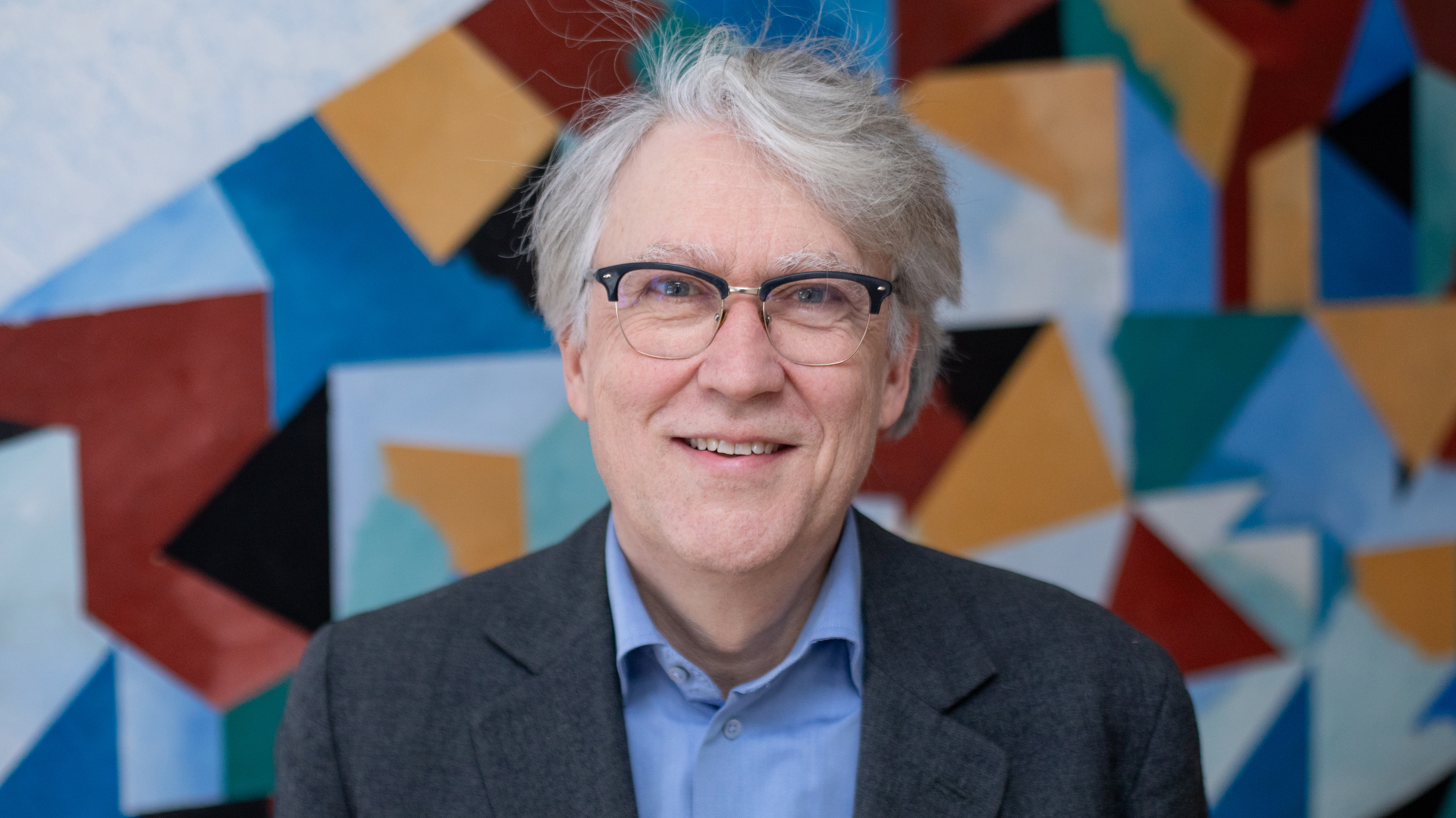 En person med grått hår och glasögon står framför en färgrik abstrakt målning
