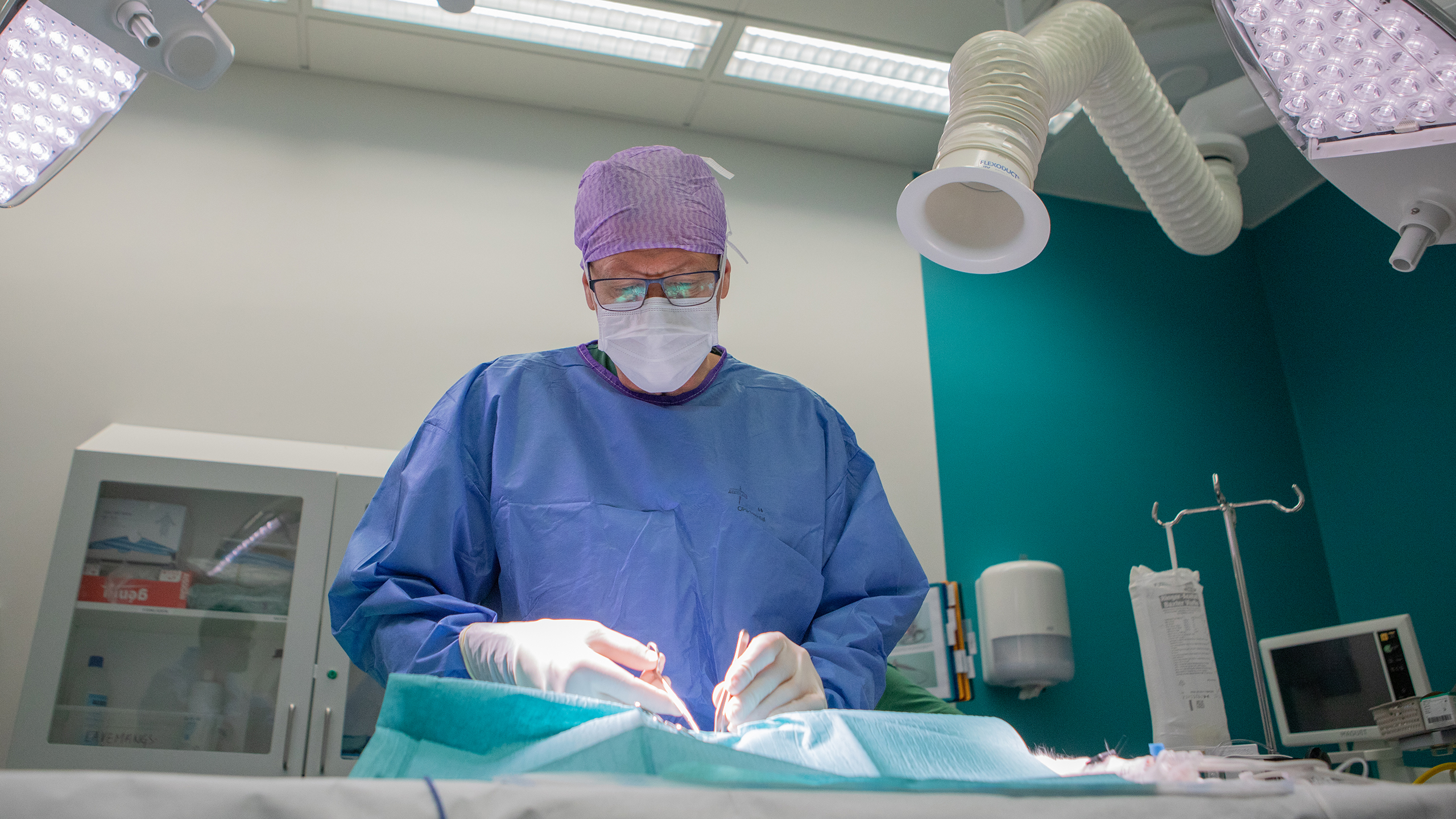En kirurg med sjukhuskläder, glasögon och munskydd står i en operationssal.