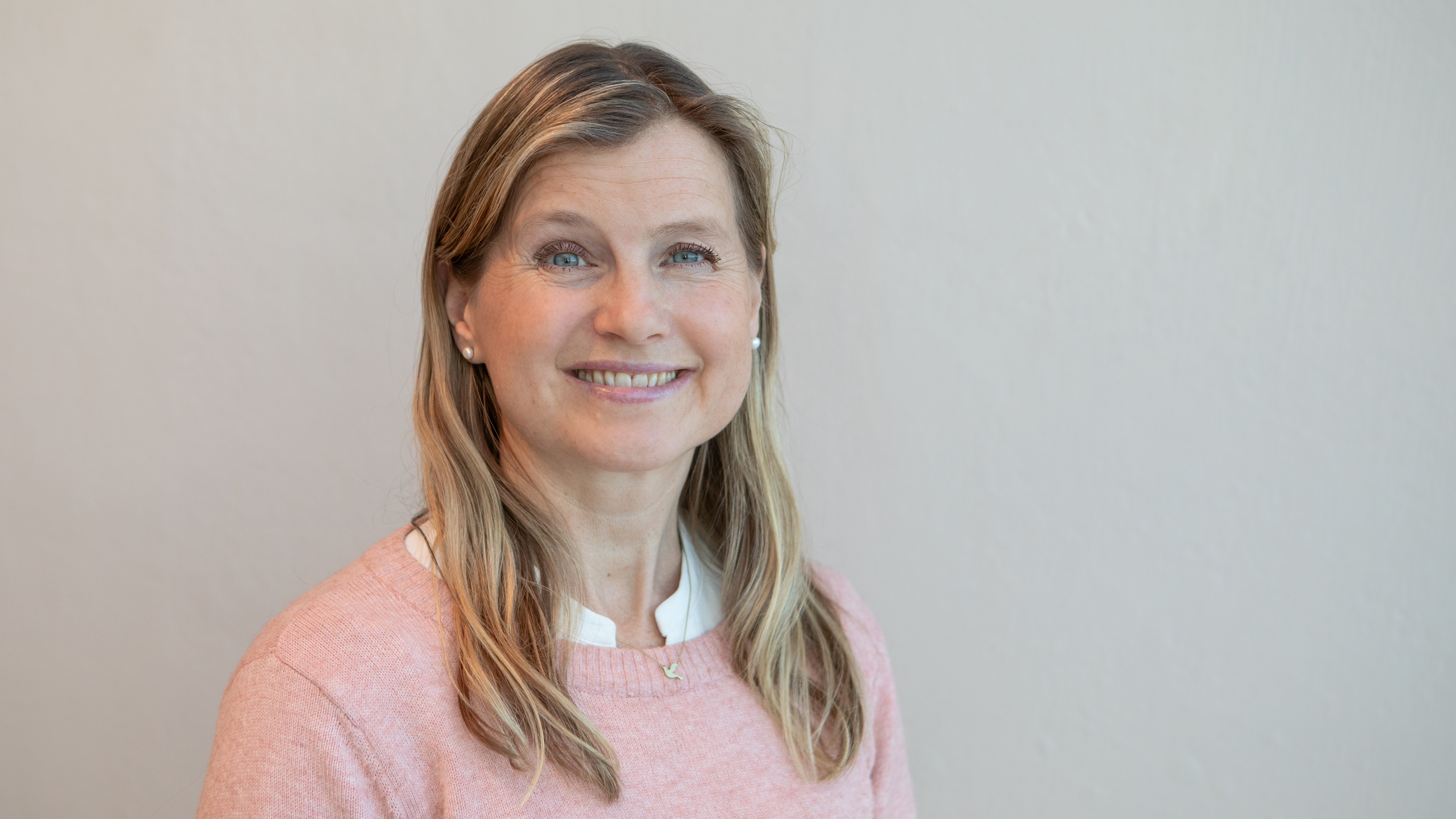 A woman with blonde hair, wearing a pale red shirt smiles into the camera. 