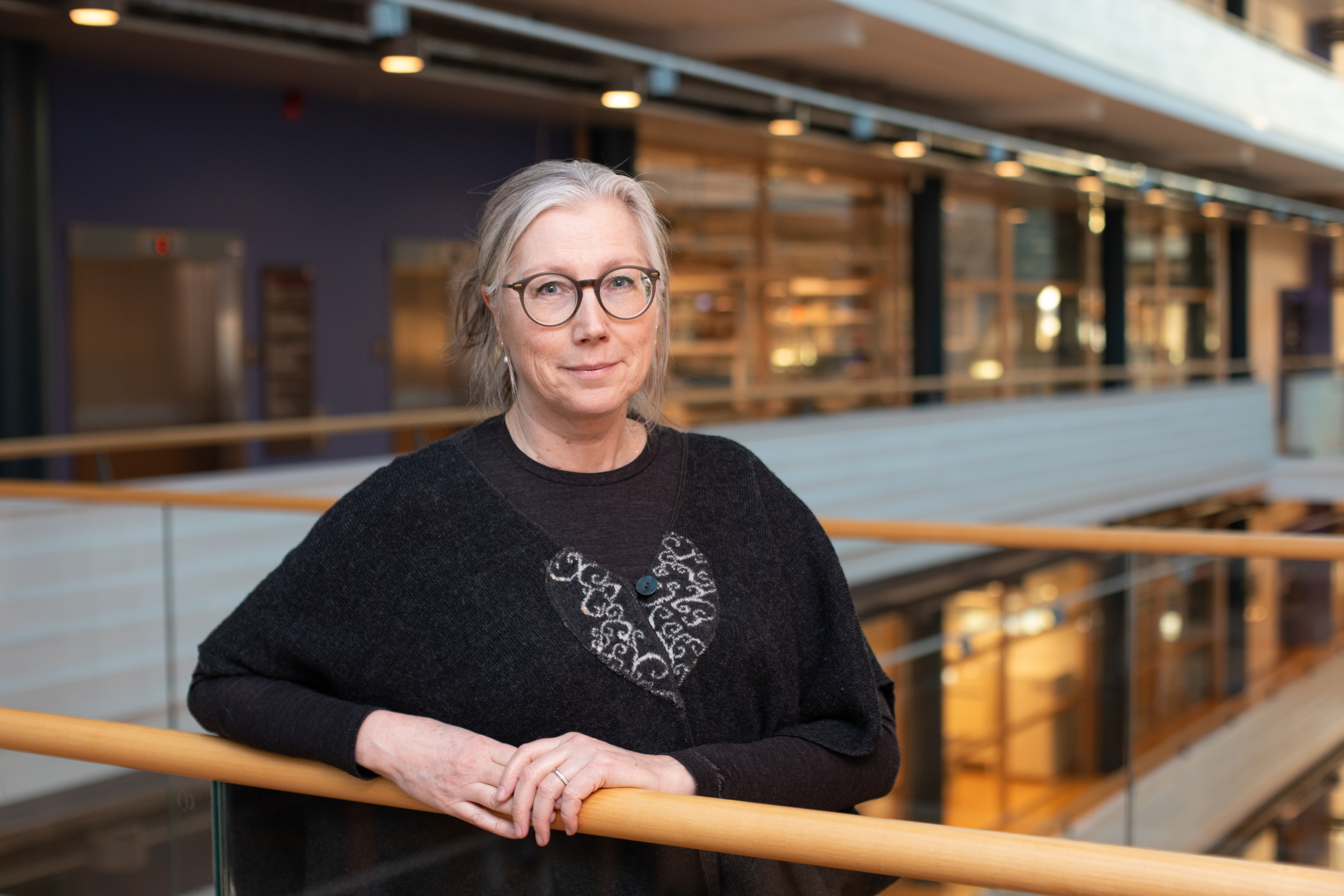 En kvinna med glasögon står i en modern stor trapphall och tittar in i kameran.