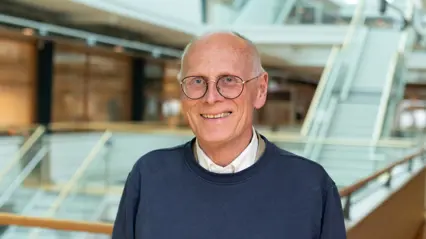 A person with grey hair and wearing glasses, a shirt and a wool shirt smiles into the camera.