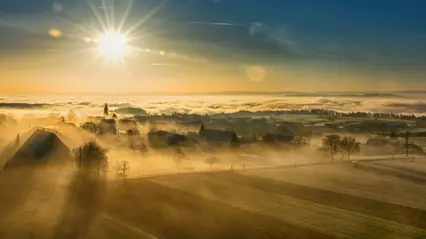 Solen lyser lågt över ett dimmigt landskap med hus och åkrar.