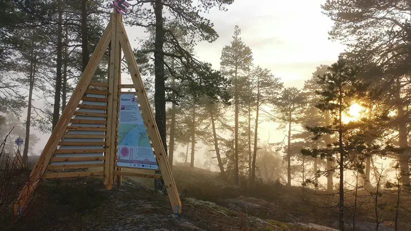 En skylt står på ett berg, solen skymtar bakom några tallar.