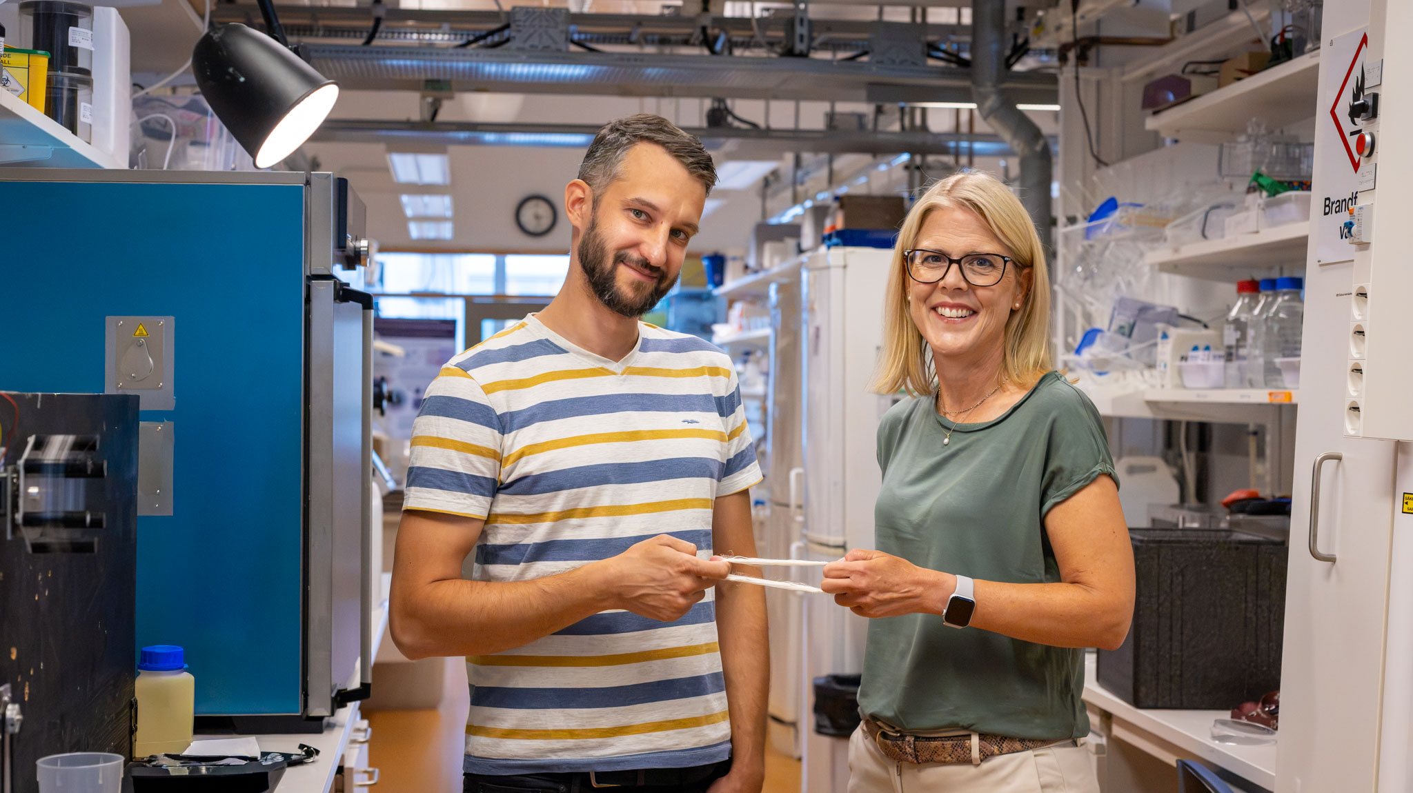 A man and a woman stand in a laboratory and hold up a thin white fibre strip between them.