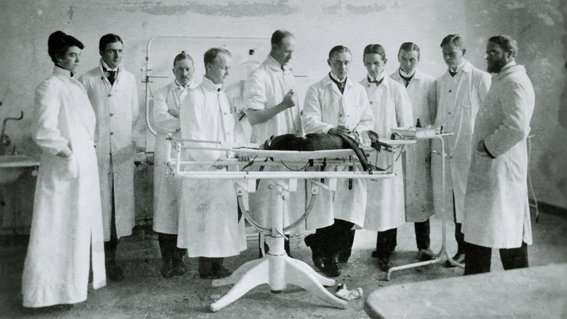 En svartvit gammal bild med en kvinna och nio män i vita rockar bakom ett djur som ligger på ett undersökningsbord.