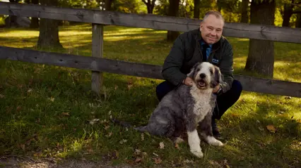A man is squatting next to his dog in a forest setting.
