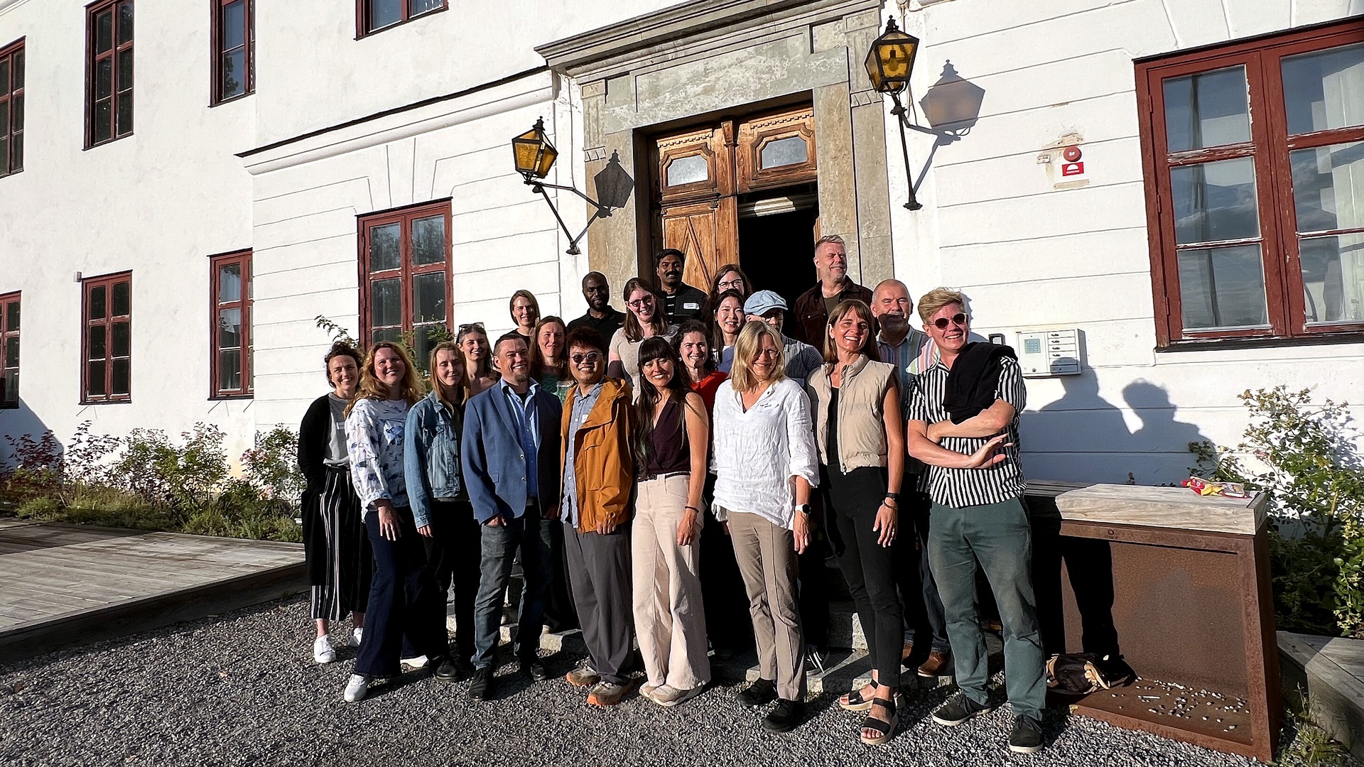 The whole group of conference attendees poses together in front of the manor's white facade and wooden door in the evening sun. 
