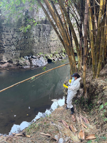 A female researcher wearing white protective gear stands by a riverbed collecting samples. Photo.