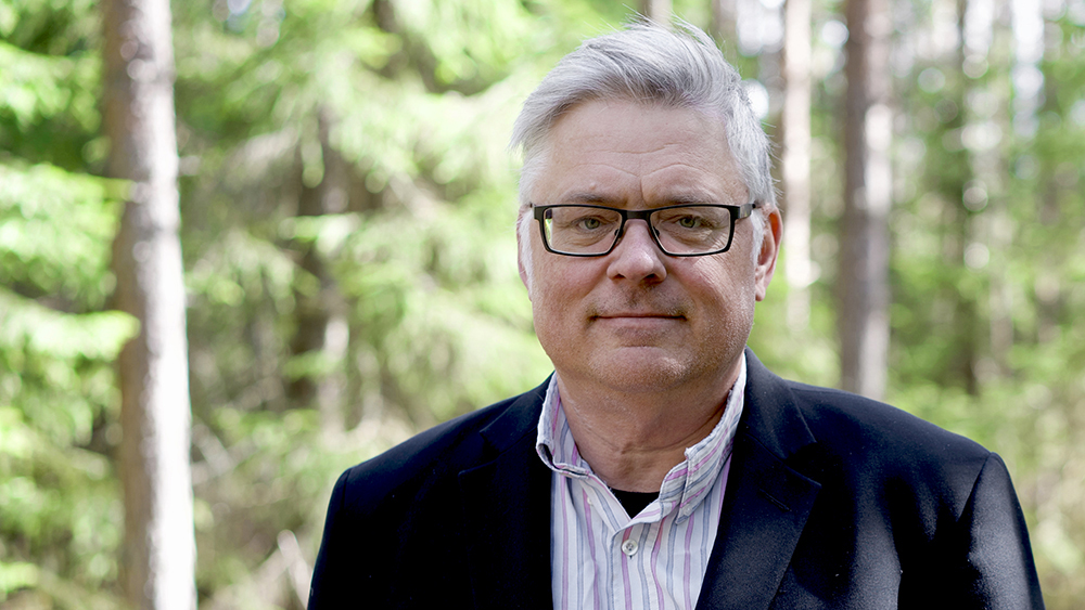 En man i mörkblå kostym och randig skjorta i en skog. Foto. 