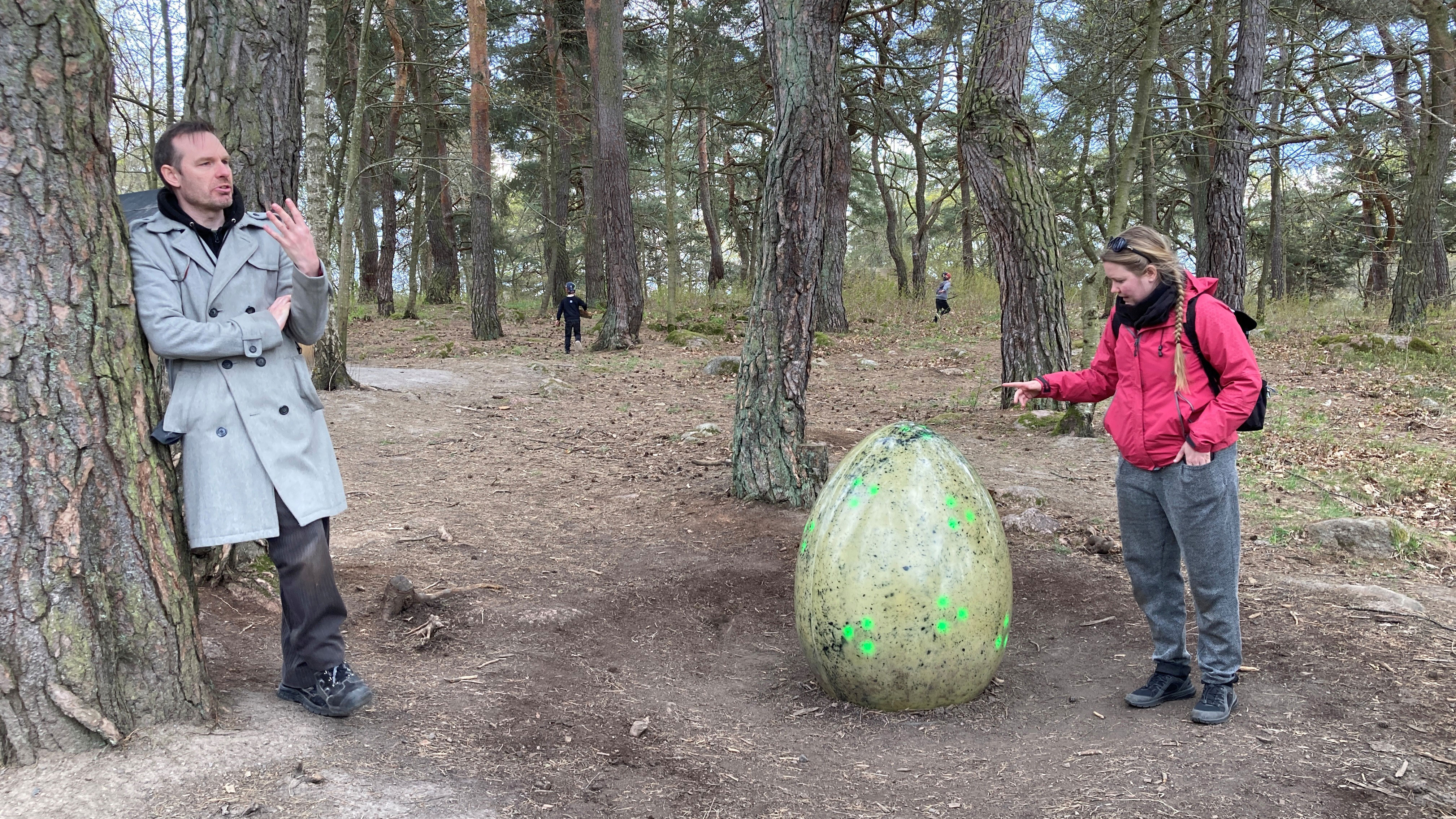 A man and a woman are in the foreground; children playing can be seen in the background. The woman is touching an egg-shaped sculpture.