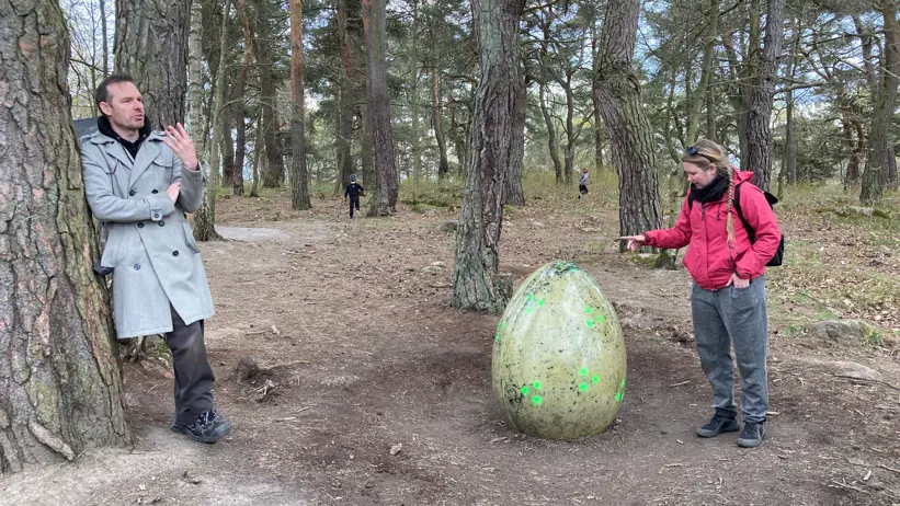 A man and a woman are in the foreground; children playing can be seen in the background. The woman is touching an egg-shaped sculpture.