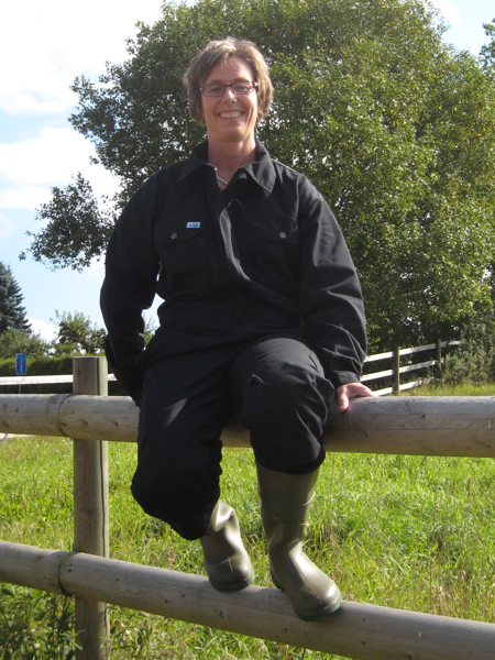 A woman in overalls is sitting on a bench in a lush outdoor setting.