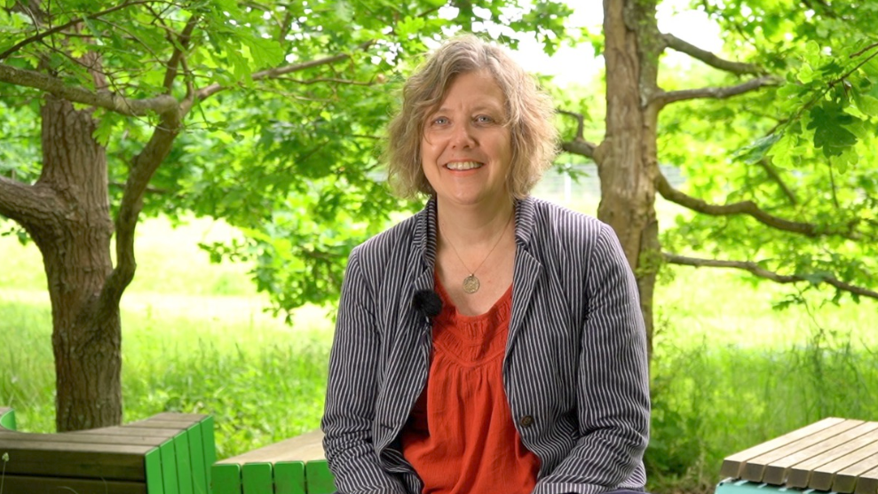 A half-length portrait of a woman in a green, summery outdoor setting.