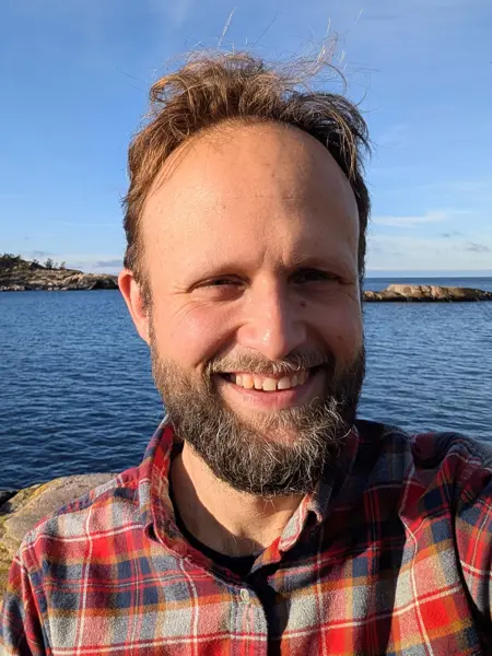 Headshot of a man wearing a plaid shirt in an outdoor setting with water in the background. Photo.