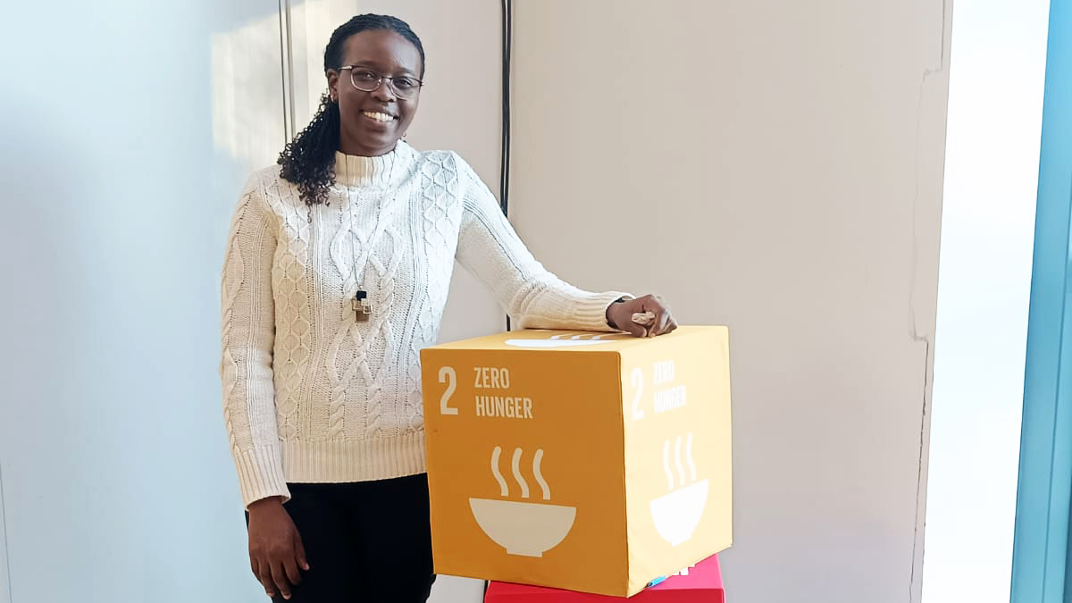 A smiling woman besides a yellow cube with the text “Zero hunger”. Photo. 