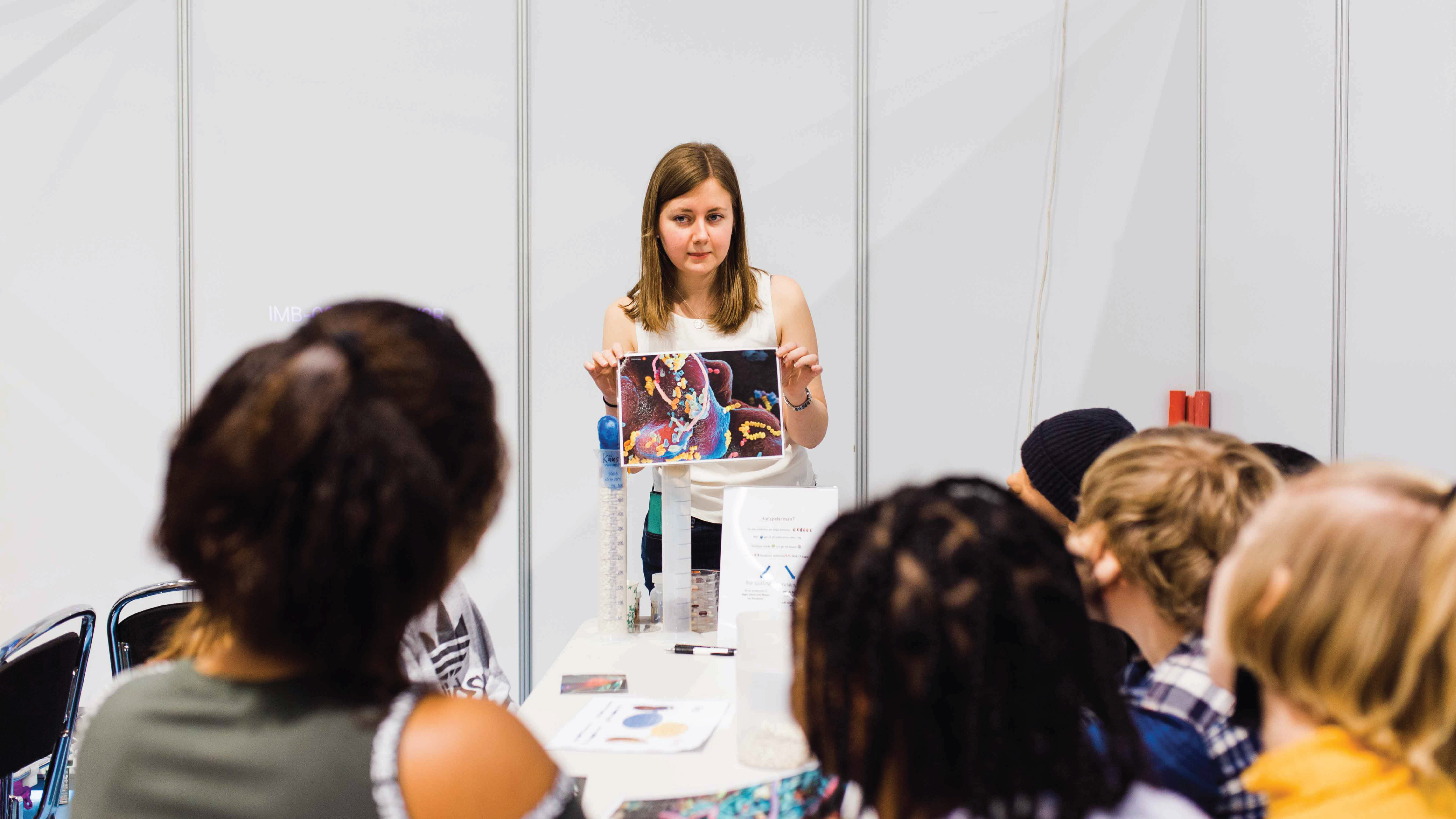 A girl shows an image in front of a small audience sitting at a table. Photo. 