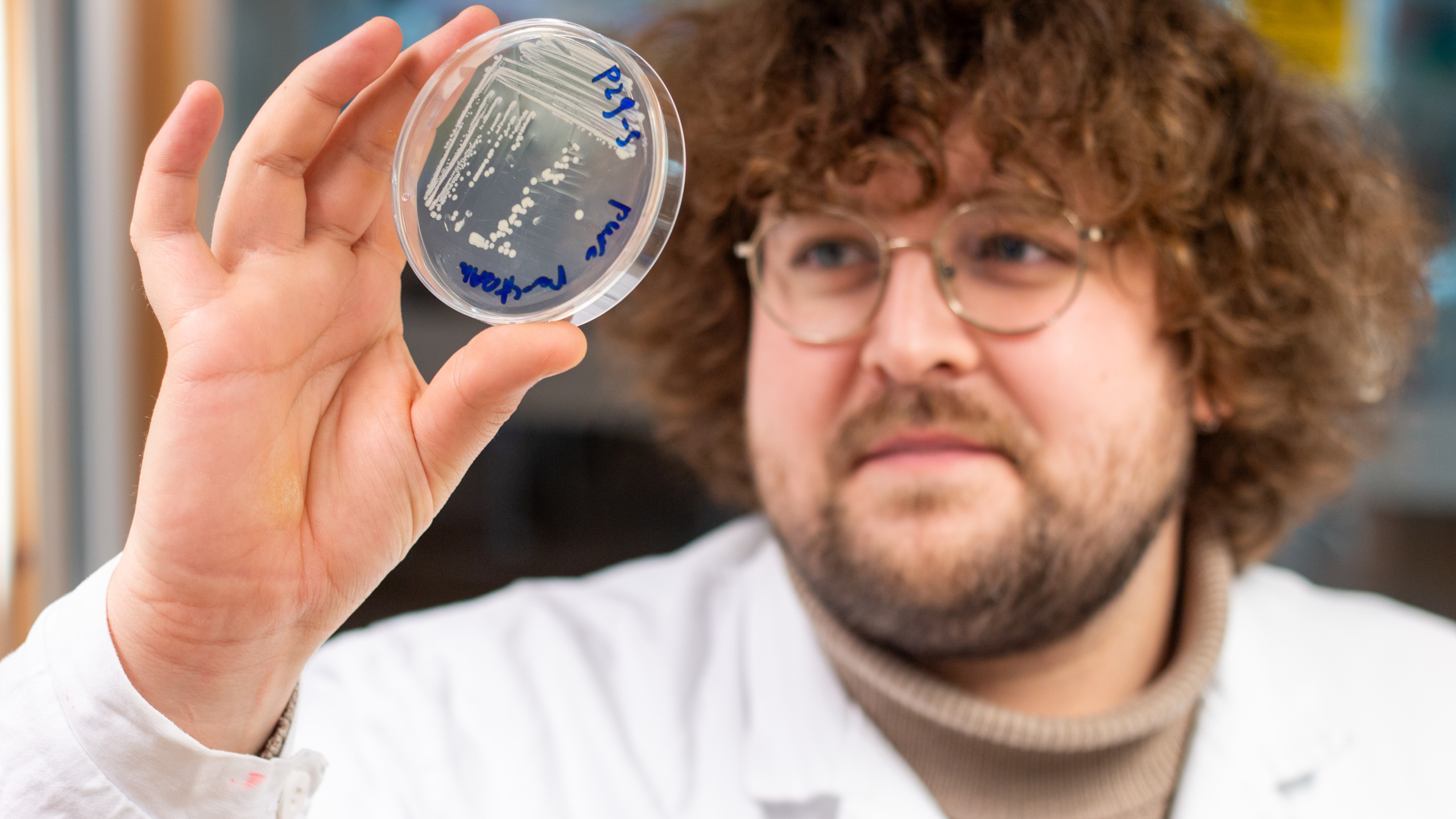 Close-up of a man in a white lab coat holding up an agar plate.