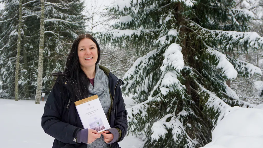 Porträttfoto av Bodil Häggström som står framför snötäckta träd, håller sin doktorsavhandling i handen och ler in i kameran. Hon har mörkt långt hår, en svart jacka och en grå tröja.