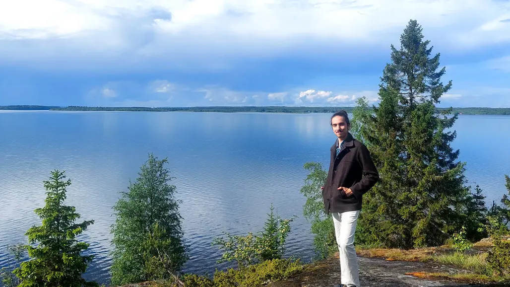 Félix Barbut wearing a white trousers and a dark jacket is standing in front of a lake in a typical Northern Swedish landscape