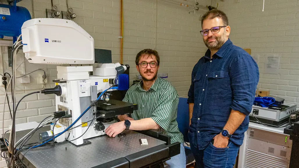 Two men at a huge microscope, one is sitting in front and one is standing next to him, both are looking into the camera.