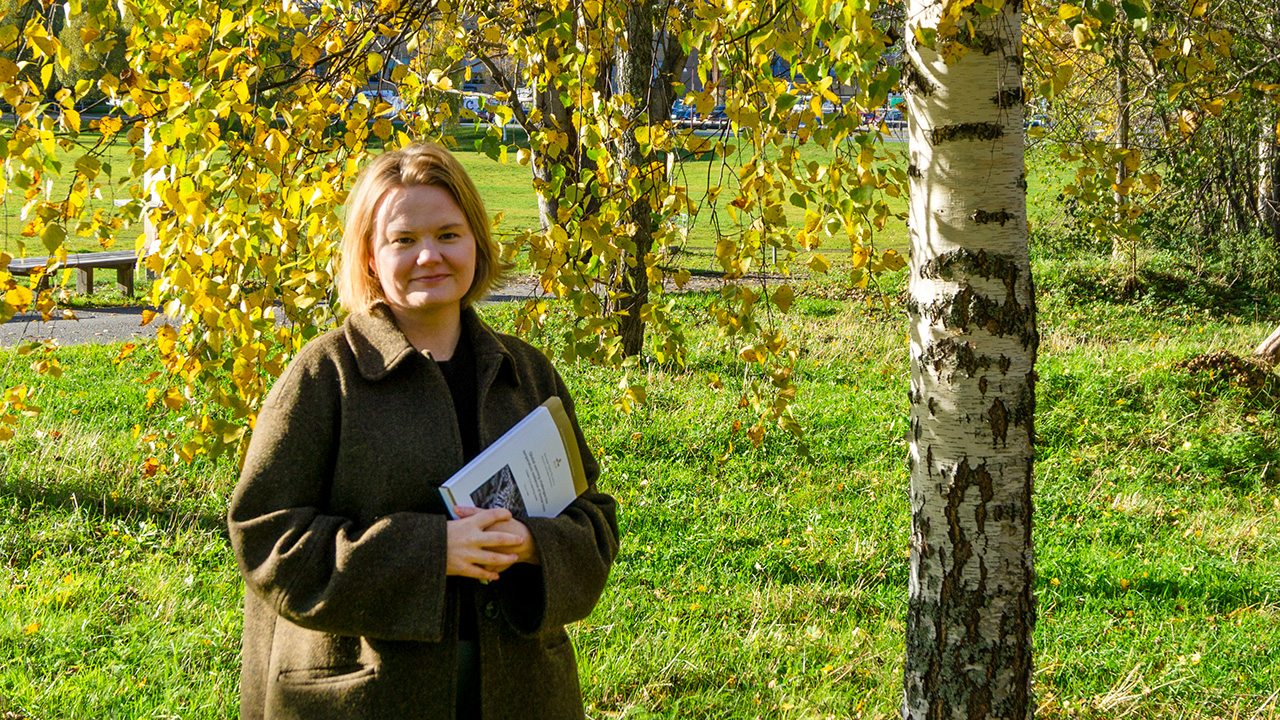 A blond women is standing next to a birch tree with yellow leaves wearing a brown coat and holding a booklet in her hands.