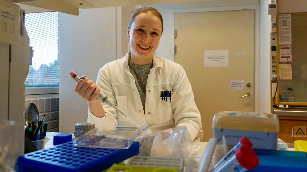 En kvinna i labbrock sitter i ett laboratorium med en pipett i handen. Hennes blonda hår är uppsatt.