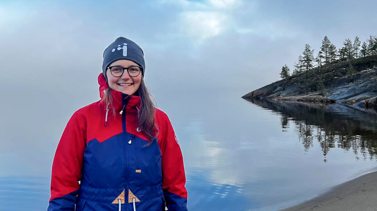 A women with glasses and long brown hair is standing at a water front. She is wearing a hat and a warm jacket.