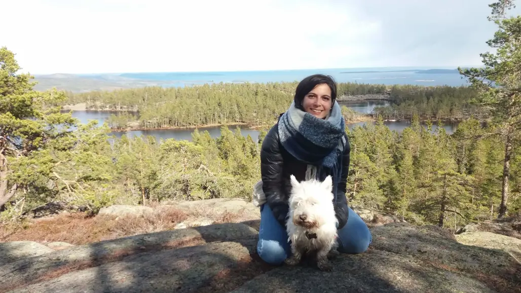 Alice Marcon is sitting on a stony hill in front of a forest landscape that goes over to the sea. In front of her is sitting a small white dog. She has black hair, a scarf around her neck, jeans and a jacket on and is smiling into the camera.