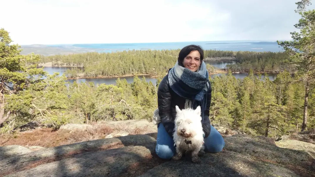 Alice Marcon is sitting on a stony hill in front of a forest landscape that goes over to the sea. In front of her is sitting a small white dog. She has black hair, a scarf around her neck, jeans and a jacket on and is smiling into the camera.
