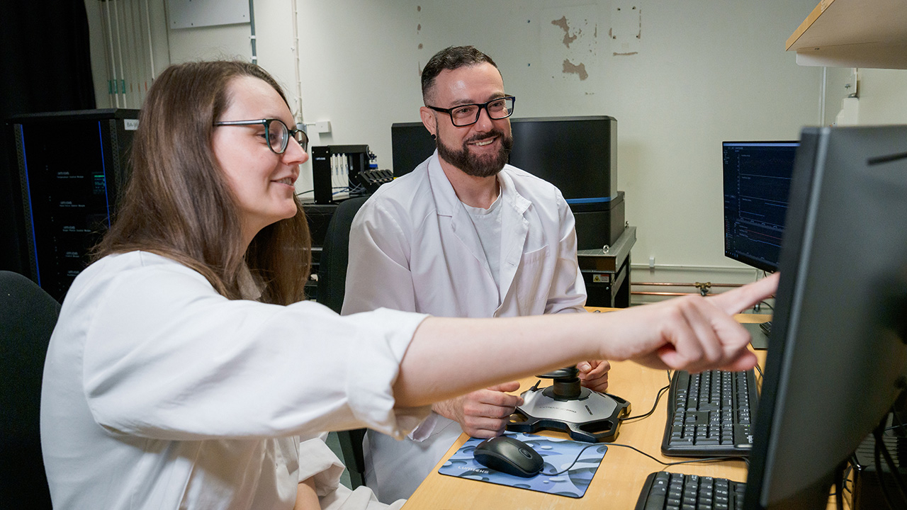 A women and a men in white labcoats are sitting in front of a computer screen and discuss. The women is shown on something on the screen.