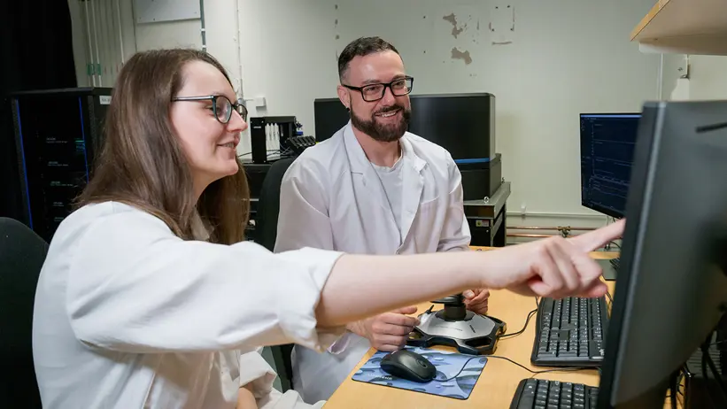 A women and a men in white labcoats are sitting in front of a computer screen and discuss. The women is shown on something on the screen.