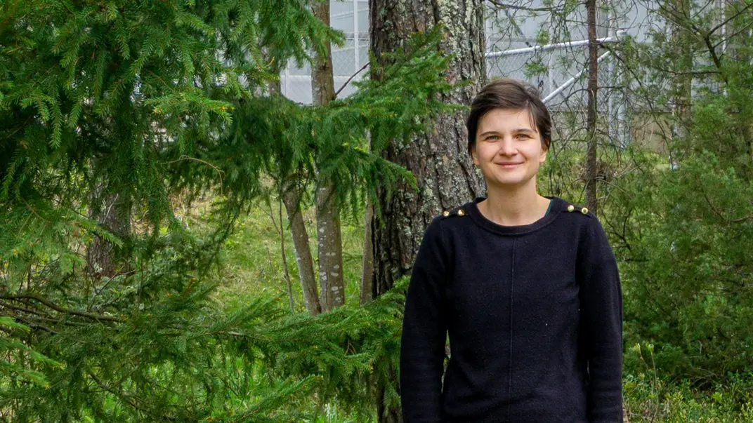 Portrait of Tinkara Bizjak-Johansson who has short dark hair and is wearing a black sweater. She stands outside next to a pine tree with a greenhouse in the back.