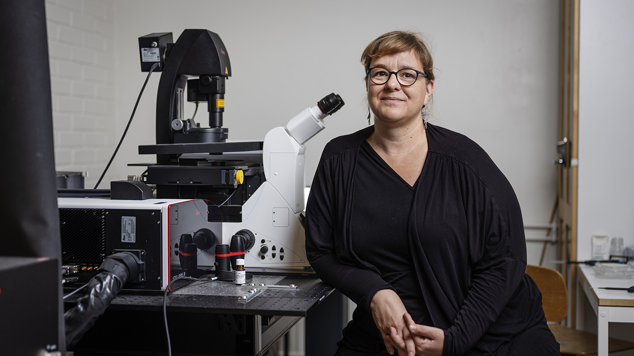 Portrait of a woman with long brown hair and black clothes who is sitting next to a microscope