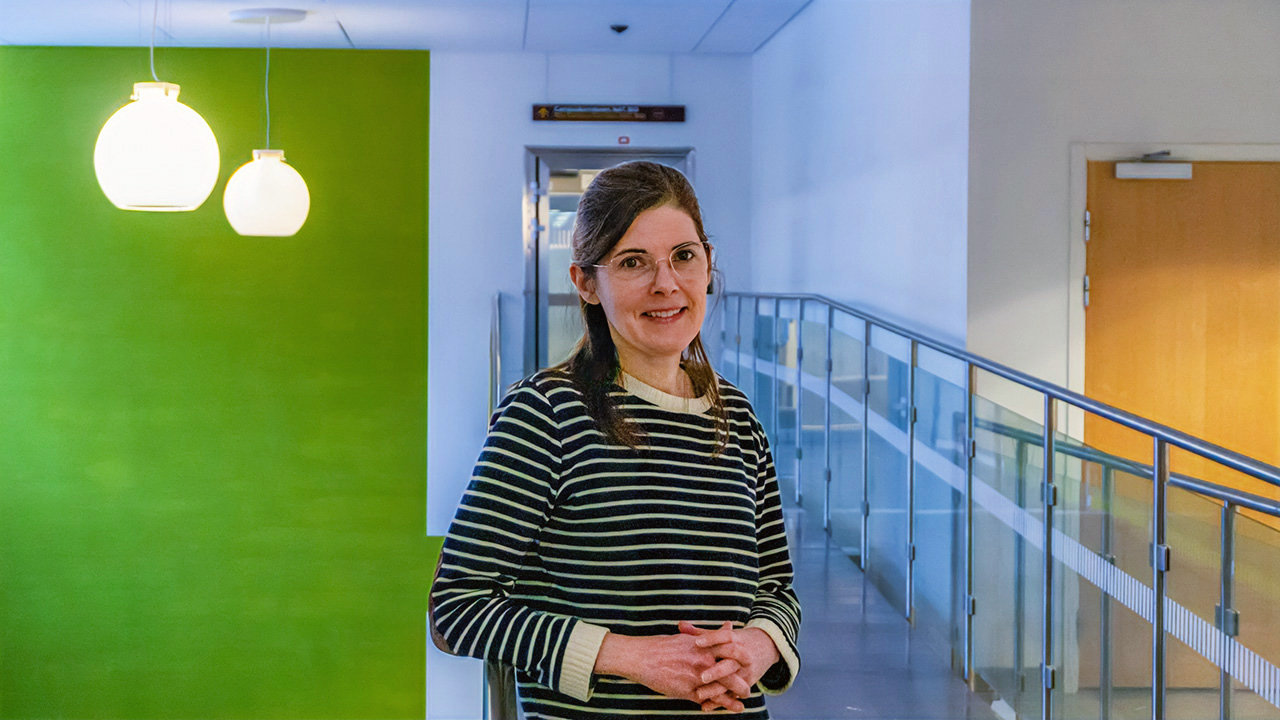 Susan McEvoy, klädd i en randig tröja och glasögon, står på en gångbro inomhus. I bakgrunden till vänster syns en grön vägg.