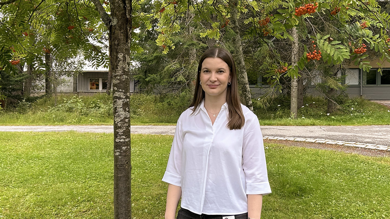 Julie Ducla står framför ett träd med röda bär och tittar in i kameran. Hon har på sig en vit blus.