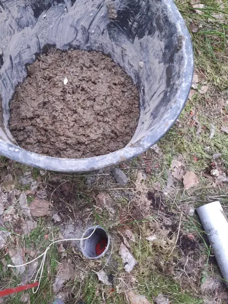 En hink med jord som står på skogsmarken bredvid ett litet hål i marken och ett metallrör
